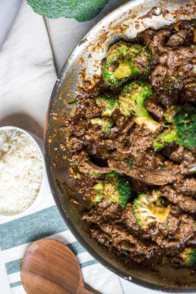 a skillet full of beef and broccoli with a small bowl of white rice in the upper right corner