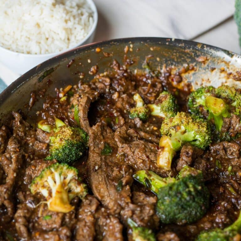 a skillet full of beef and broccoli with a small bowl of white rice in the upper left corner