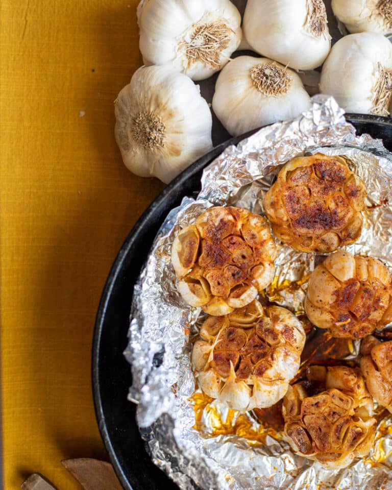 five oven roasted garlic bulbs in a cast iron pan with foil with six raw whole garlic bulbs on the side of the cast iron