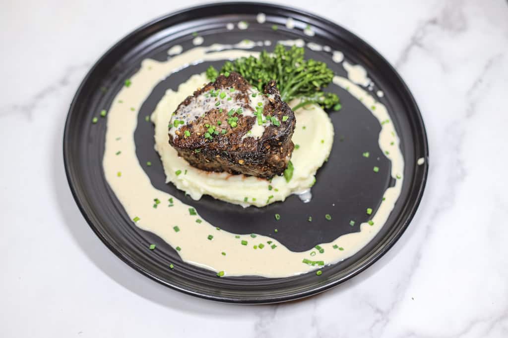 filet mignon plated over a bed of mashed potatoes with a creamy peppercorn sauce drizzled on top