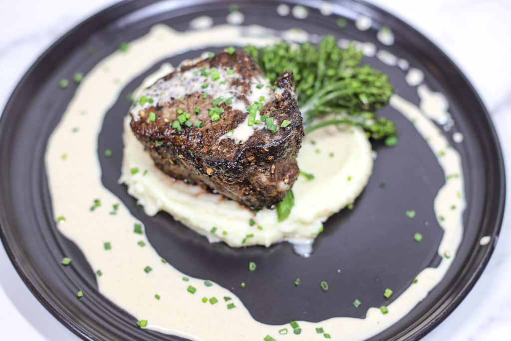 filet mignon plated over a bed of mashed potatoes with a creamy peppercorn sauce drizzled on top