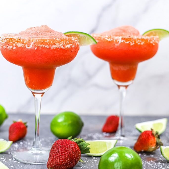 Two strawberry frozen margaritas with tajin, salt, and lime rim. Limes and strawberries around bottom of glasses