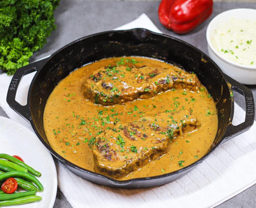 smothered pork chops in cast iron topped with parsley. parsley, green beans, bell pepper, and mashed potatoes in background