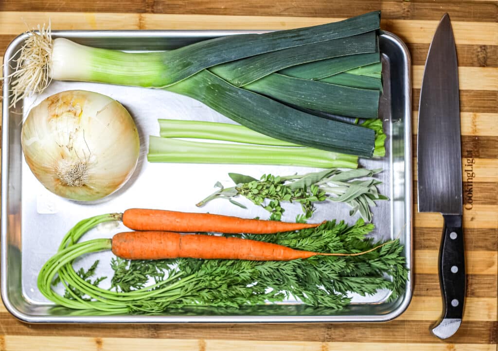 white onion, carrots, leeks, celery, dried thyme, and parsley on silver pan. knife on right side of pan