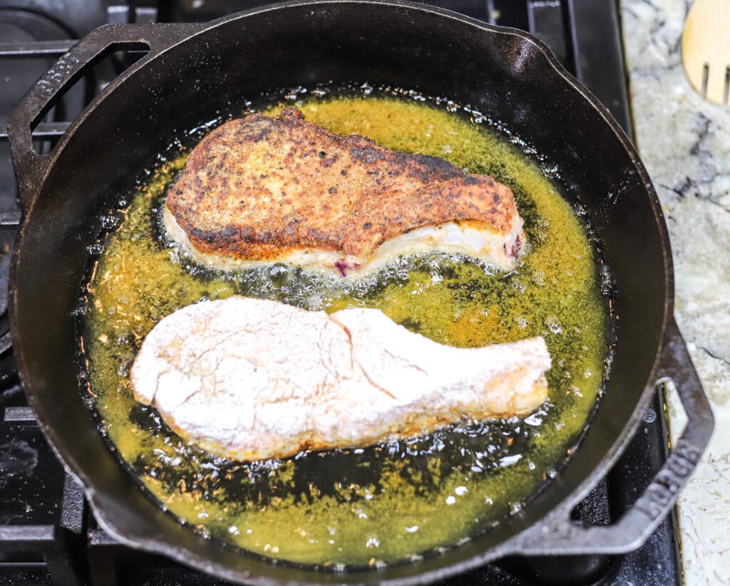 Two pork chops cooking in oiled cast iron skillet. One pork chop is shown with fried side up